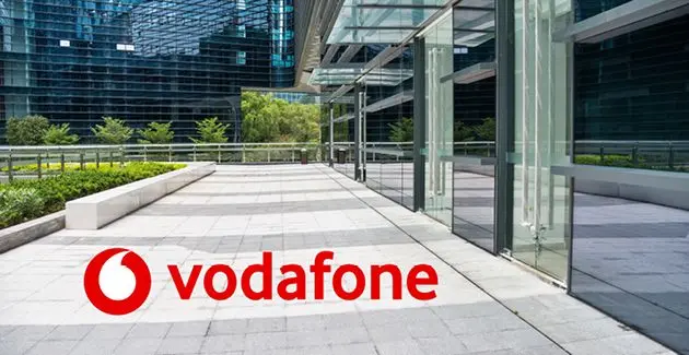 The red and white Vodafone logo superimposed on a paved outdoor walkway in front of a modern glass office building.