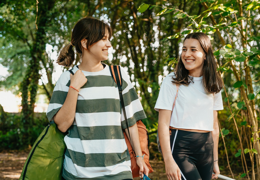 Two young women smiling and walking together outdoors in a green, leafy area. One carries a rolled sleeping bag and the other a backpack, suggesting they are on a camping or hiking trip. Sunlight filters through the trees.