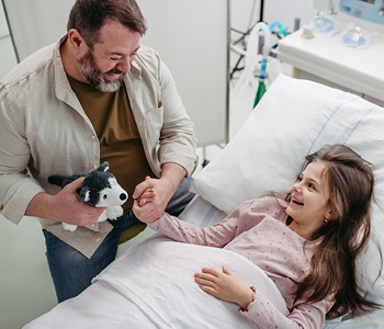 Srečen trenutek v bolnišnici, deklica leži v postelji, oče pa jo drži za roko, vesel okrevanja. Oba se smejita.