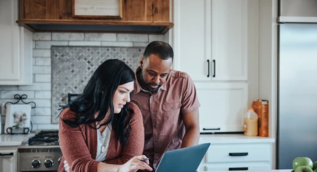 Man and woman sit at kitchen counter with breakfast working with pen, paper and laptop.    