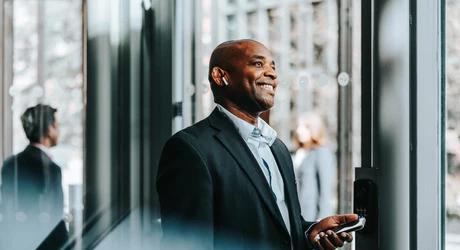 Person standing by a glass door in an office building, holding a smartphone and wearing wireless earbuds. 