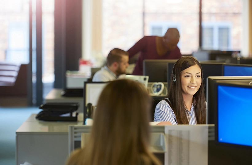 Photo of female Contact centre support worker with headset on at her desk talking to someone. Office environment