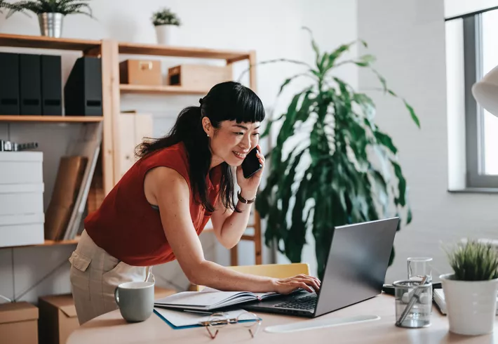 Professional Asian woman on phone leaning over desk typing on laptop in small office 