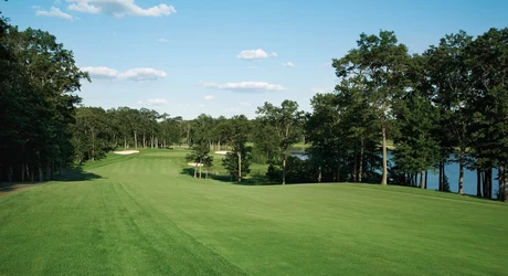 Well-maintained golf course fairway and green surrounded by trees, with a lake and blue sky in the background.
