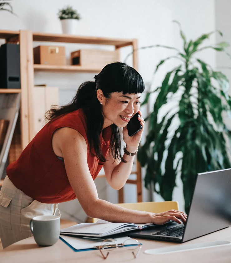 Professional Asian woman on phone leaning over desk typing on laptop in small office 