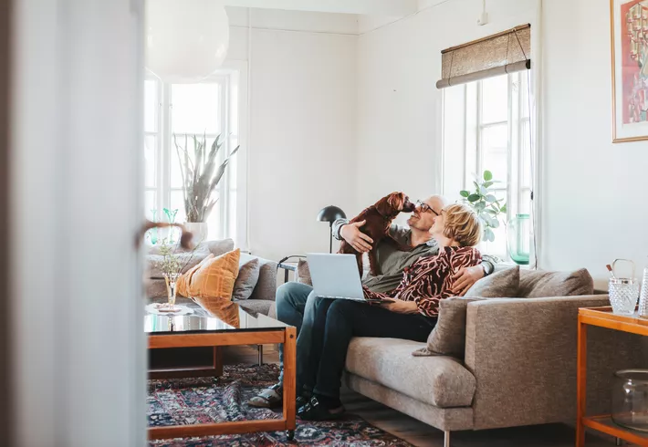 A couple sitting on their sofa in the living room, with a laptop and their dog. 