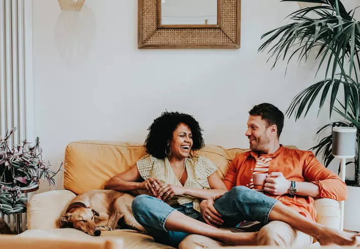 Two adults sit on a couch in a cozy living room, holding mugs and talking while a dog sleeps beside them; houseplants, framed wall art, and a floor lamp decorate the space. 