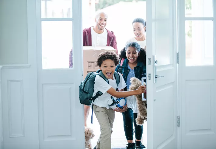 Two children enter through an open front door into a home, one holding a backpack and sports ball and the other carrying a stuffed toy, while two adults follow behind holding a moving box; a small dog is partially visible near the doorway. 