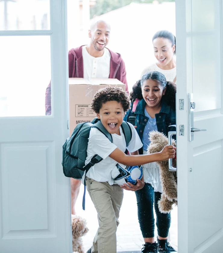 Two children enter through an open front door into a home, one holding a backpack and sports ball and the other carrying a stuffed toy, while two adults follow behind holding a moving box; a small dog is partially visible near the doorway. 