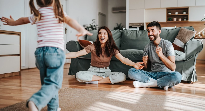  Child running toward two adults sitting on the floor of a living room, with a couch and kitchen visible in the background. 