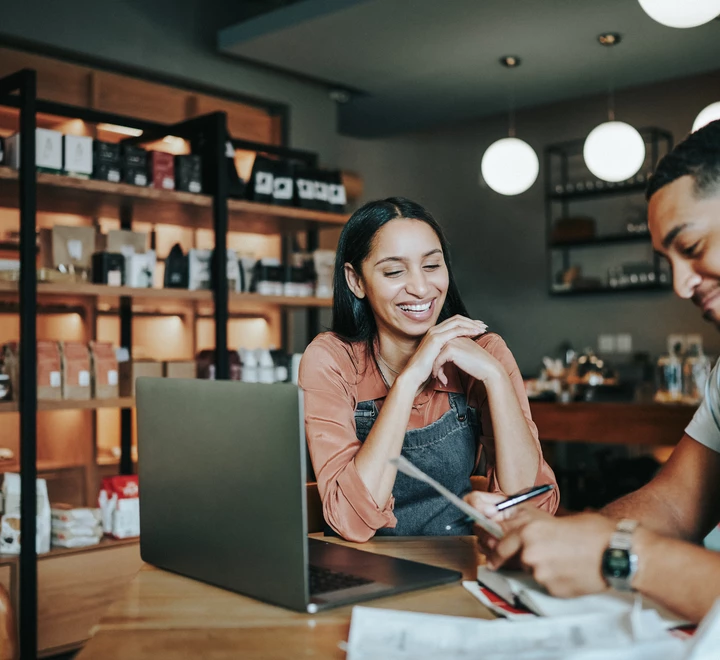 Two smiling young delicatessen owners going over paperwork and working on a laptop at a table in their artisanal shop 