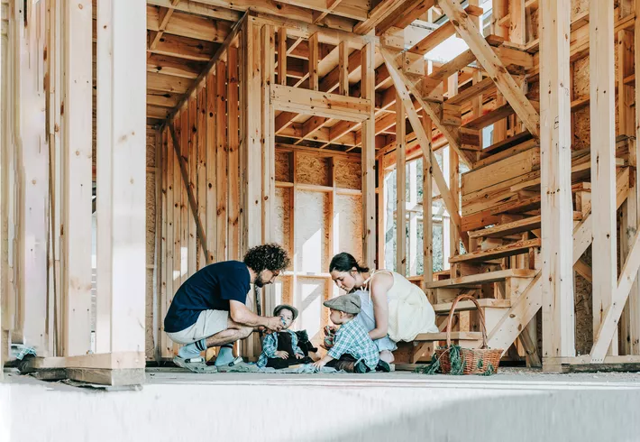 Two adults crouch beside two young children on the floor of a house under construction, surrounded by exposed wooden framing and a staircase, as they gather in the unfinished interior. 
