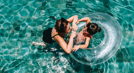 Mother floating in a pool gently guides her young daughter, who is in a water ring, through the sparkling, tiled pool. 