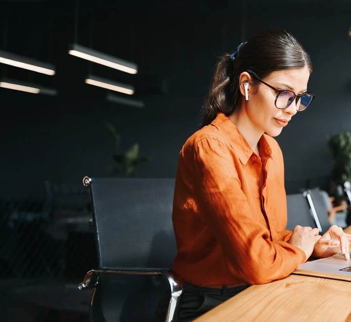 Professional woman working on a laptop, sitting at a desk next to a window in an office setting. 