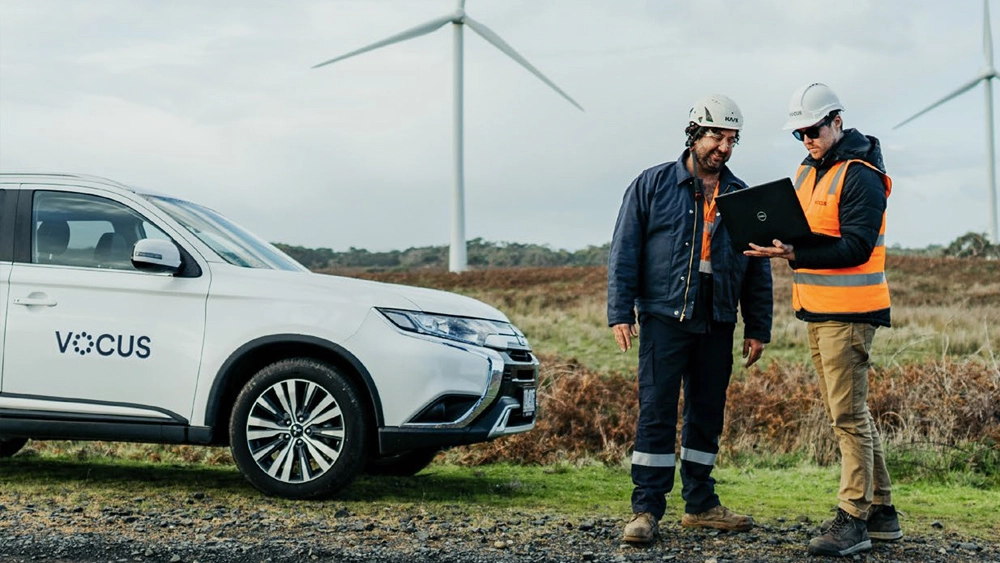 Image taken from the cover of the Vocus Whitepaper: Connectivity 4.0: Achieving Sustainable Growth. Is a picture of two workers in hard hats looking at plans. A wind farm is in the background with a Vocus logoed car.